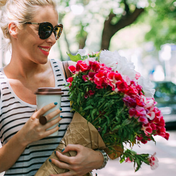 A happy woman on a city street holding a bouquet of pink flowers and a coffee